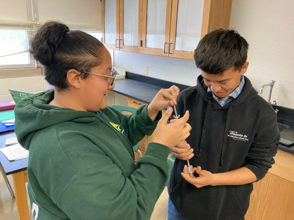 Two students in a classroom work together to measure a metal tool during a hands-on science activity.