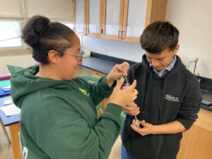 Two students in a classroom work together to measure a metal tool during a hands-on science activity.