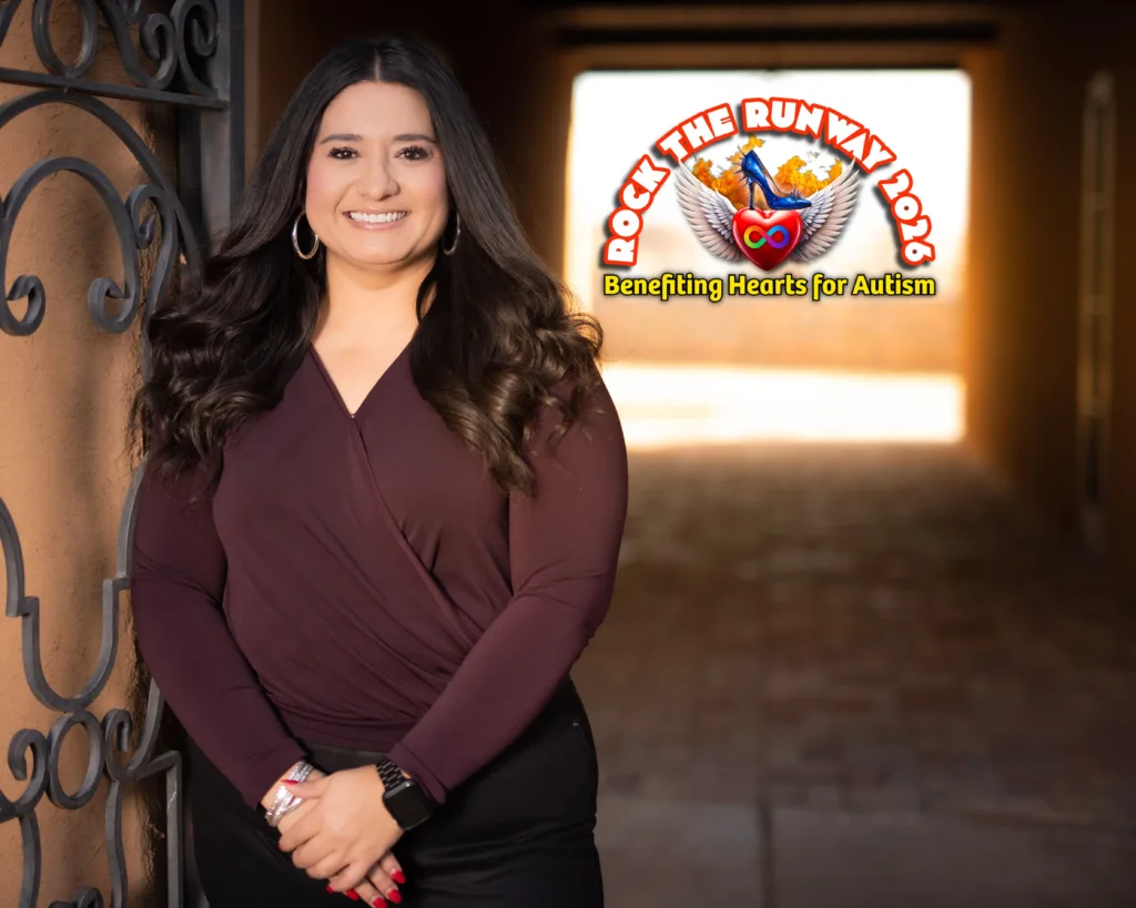 Smiling Krystal in maroon blouse posing beside an iron gate with Rock the Runway 2026 Benefiting Hearts for Autism logo in the background